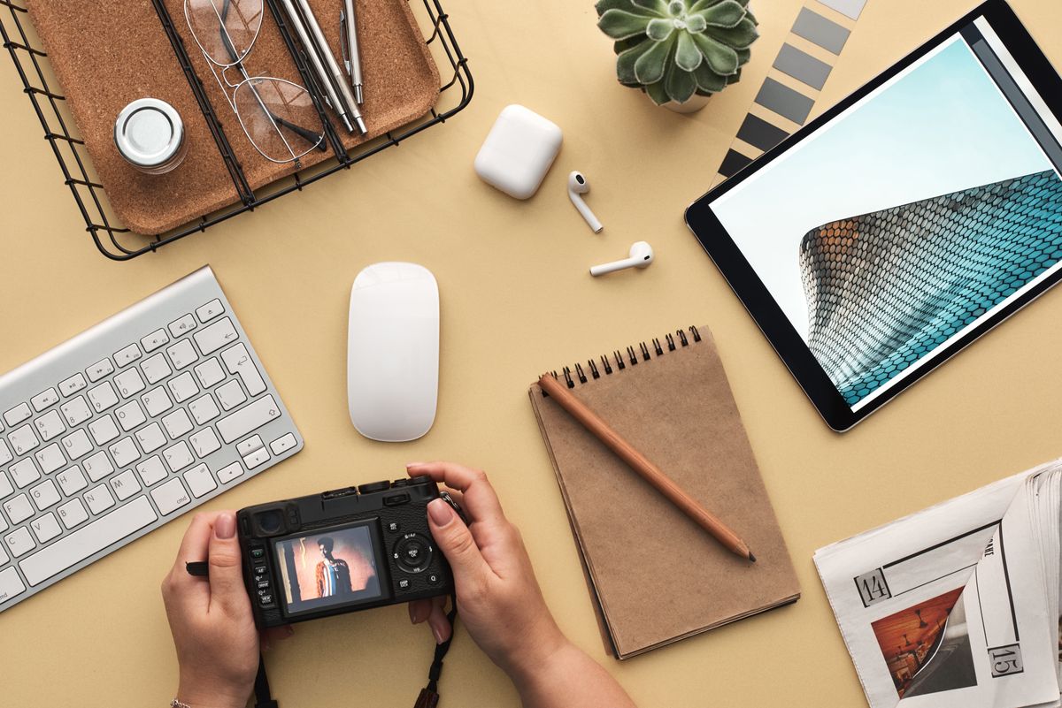 Schreibtisch von oben mit Kamera, Tablet, Tastatur, Maus, Notizbuch und Pflanze im Hellbeige-Ton